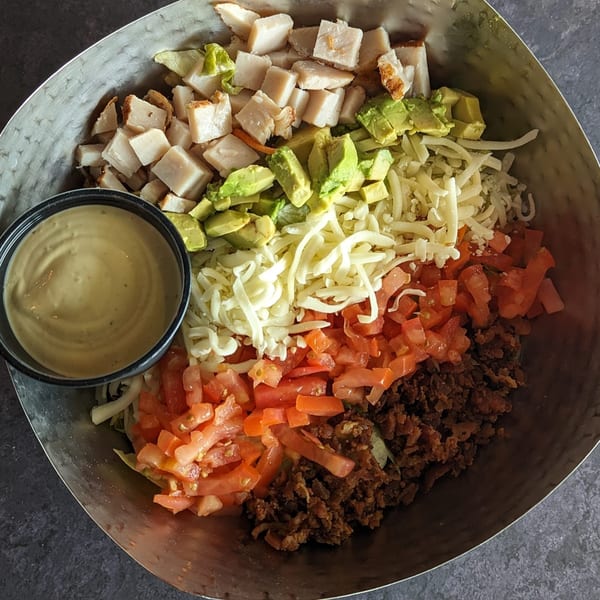 A large metal bowl of Cobb salad with chicken, avocado, cheese, tomatoes, and bacon, with dressing.