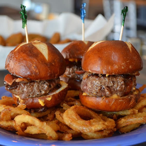 Three Rep's Place pretzel bun sliders with bacon and cheese, surrounded by crispy onion rings.