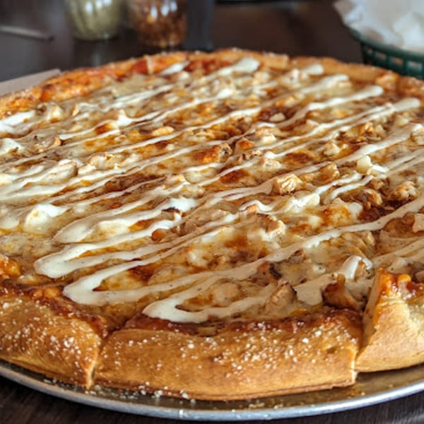 A whole Rep's Place pizza with chicken and white sauce drizzled on top, on a metal tray.