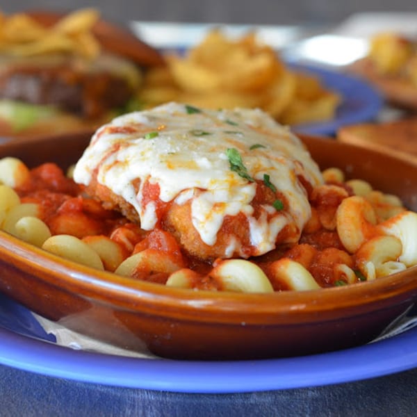 Chicken parmesan with pasta in a brown bowl, with other dishes blurred in the background at Rep's Place.